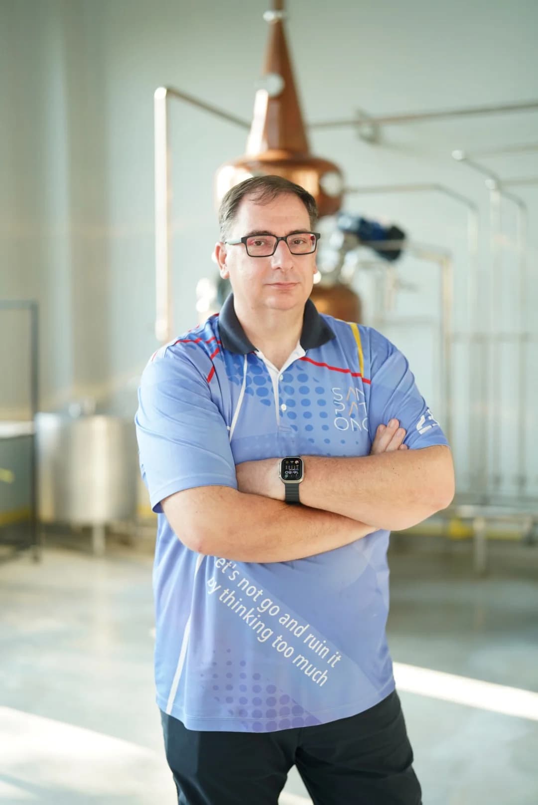 Stefan Rakoczi สวมแว่น ใส่เสื้อโปโลสีน้ำเงิน ยืนกอดอกในโรงกลั่น มีหม้อต้มทองแดงและถังสแตนเลสด้านหลัง