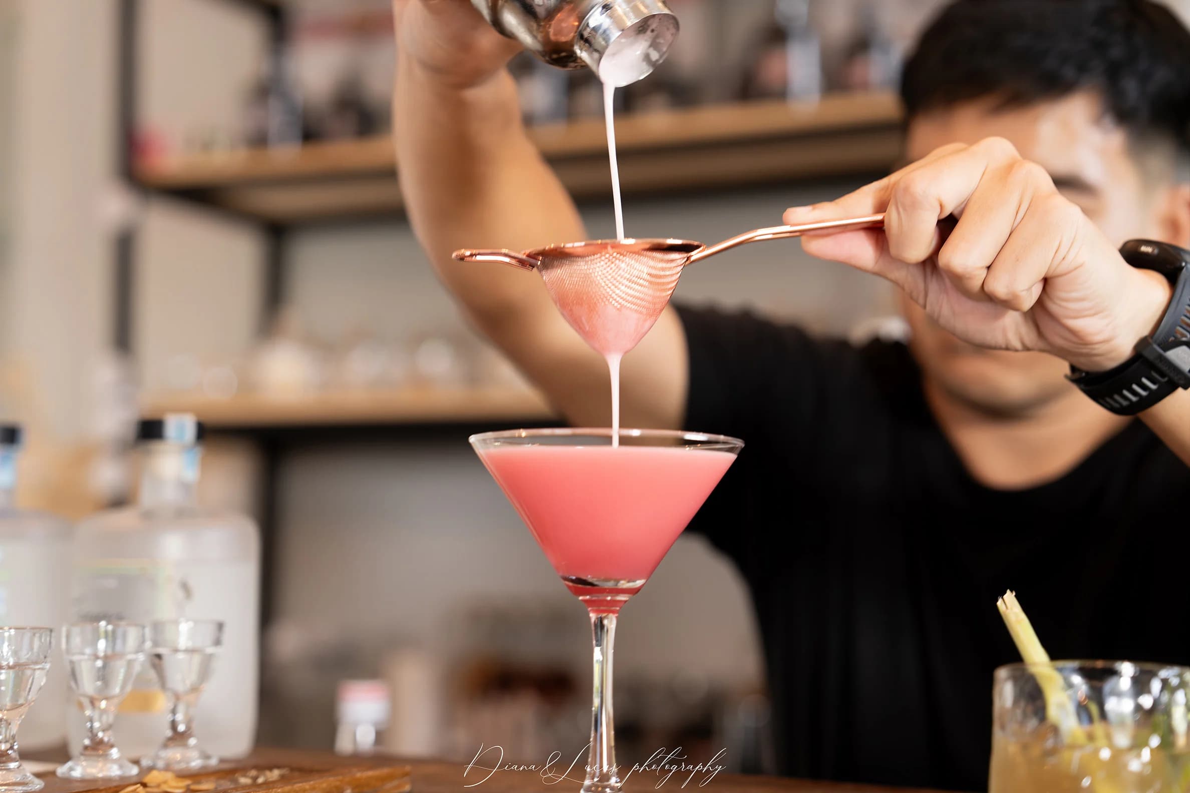 Bartender strains a pink cocktail into a martini glass at a bar, pouring liquid through a metal strainer during drink preparation.