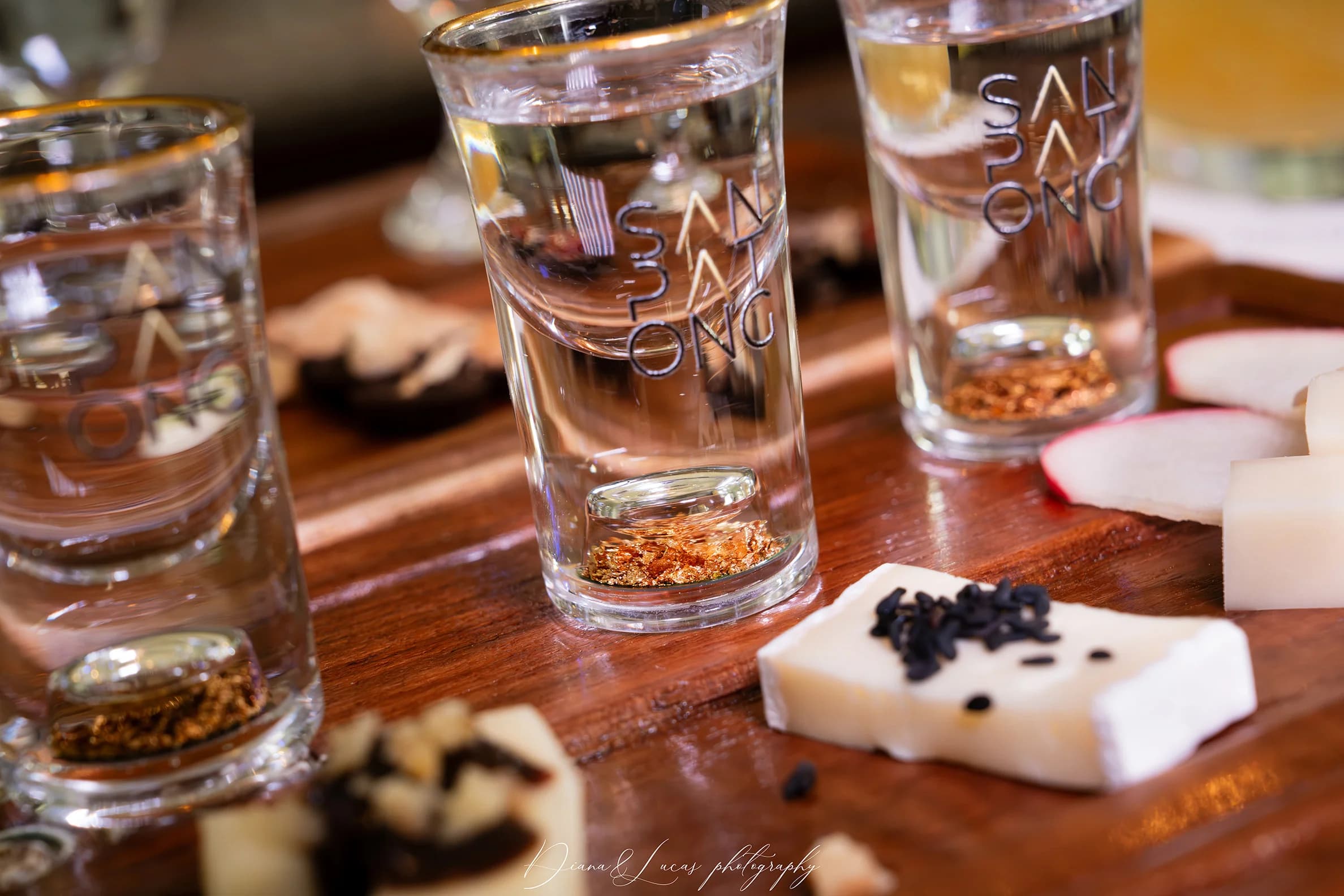 Close-up of Sanpatong shot glasses with infused spirits, spices, and small tasting plates on a wooden tray.